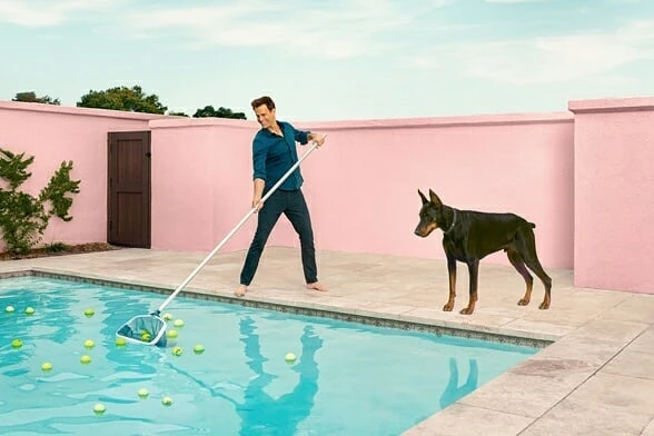 An adult male uses a pool skimmer to remove tennis balls from a blue swimming pool with a Doberman dog standing nearby outdoors.